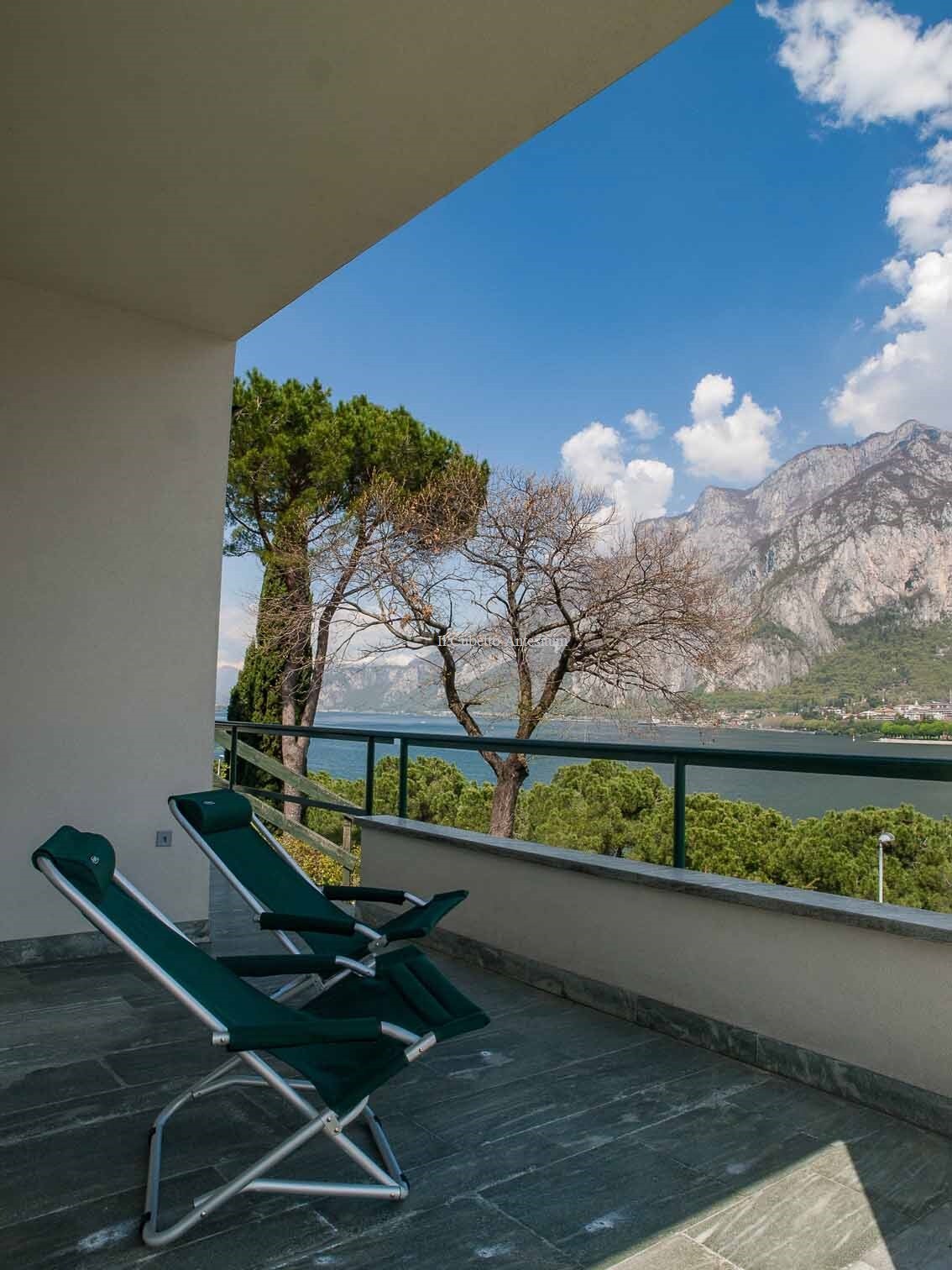 A private terrace is presented with two green reclining chairs, offering a view of Lake Como and surrounding mountains. Tall trees can be seen in the background beneath a clear sky with scattered clouds.