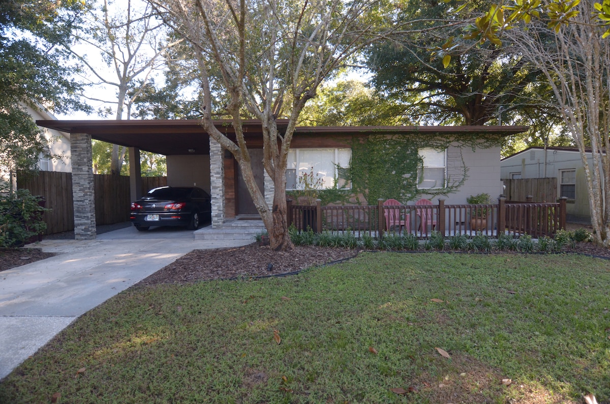 A single-story home is surrounded by lush greenery, showcasing a sloping roof supported by stone columns. A parked car is situated in the driveway alongside a wooden fence. Comfortable seating is positioned on a small patio under the shade of trees, enhancing the welcoming outdoor space.