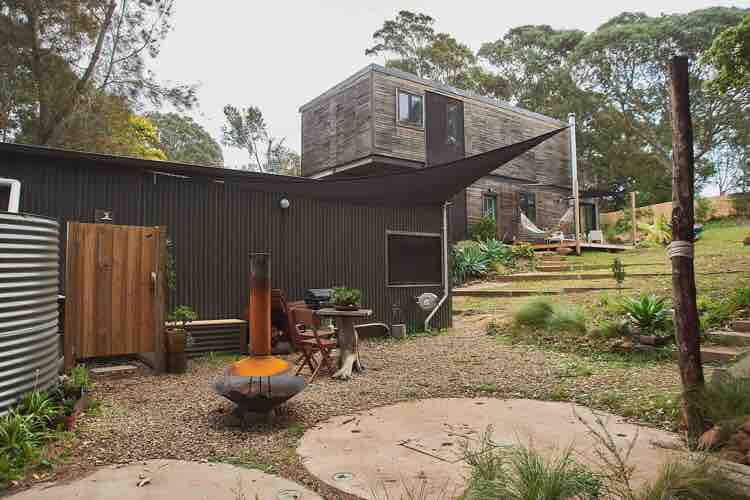 The guesthouse is surrounded by native bushland, featuring outdoor seating arrangements and a fire pit. A wooden entrance door is visible alongside a textured wall, with a shade sail providing coverage. The gardening area includes a pathway and various plants, creating a serene outdoor space.
