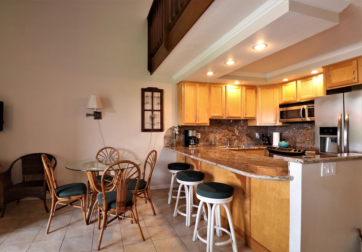 An open kitchen area features wooden cabinetry and stainless steel appliances. A kitchen island with seating for five is positioned alongside a glass dining table surrounded by four rattan chairs. Natural light illuminates the space through a nearby window.