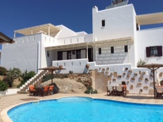 A white villa is set against a clear blue sky, featuring multiple levels and spacious terraces. A pool area is highlighted in the foreground, surrounded by lounge chairs and umbrellas. Natural stone and landscaping blend seamlessly with the architecture.