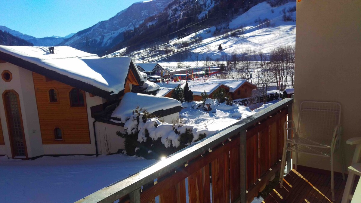 A sunny balcony offers views of snow-covered rooftops and a winter landscape. Rolling hills and distant mountains are visible, enhancing the serene atmosphere. Two chairs are positioned on the balcony, inviting relaxation in the stunning surroundings.