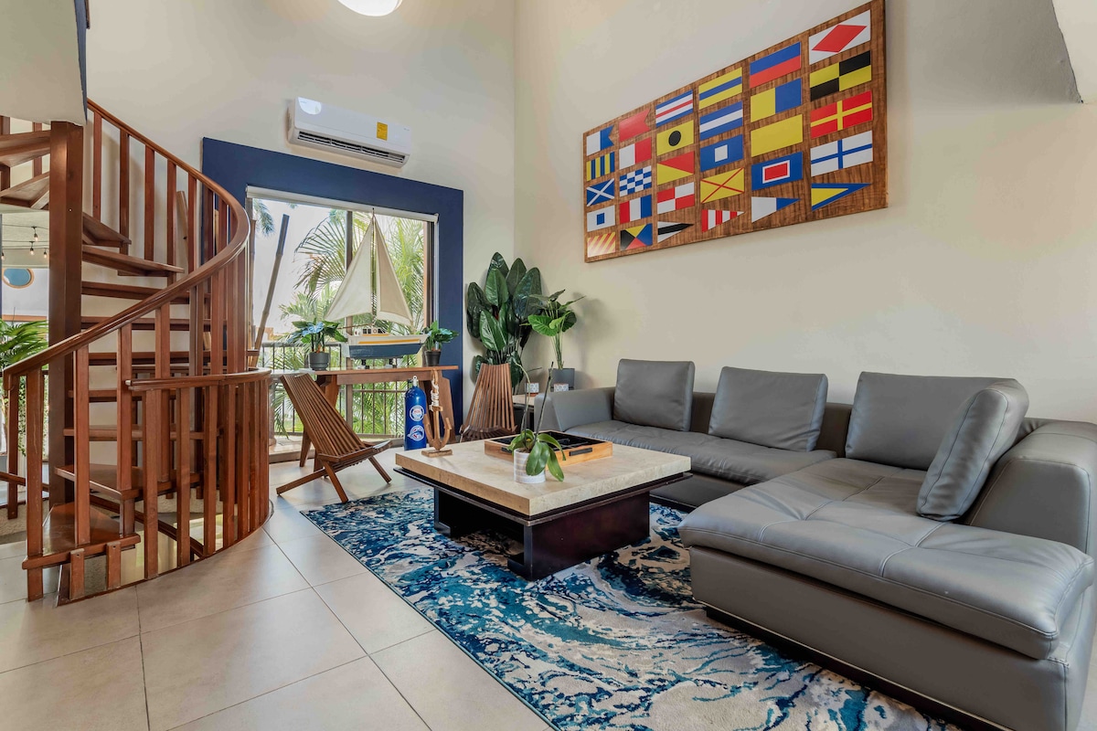 A contemporary living area features a comfortable sectional sofa and a large coffee table. Decorative elements include a vibrant rug and an artwork showcasing various flags. A spiral staircase leads to an upper level, while natural light filters through windows adorned with greenery.