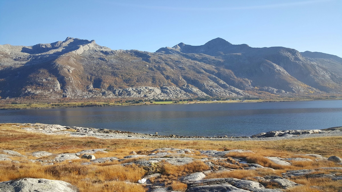 The landscape illustrates a serene lake bordered by rocky terrains and rolling mountains. Autumnal foliage adds warm tones to the scenery, while the clear blue sky enhances the peaceful ambiance. The scene invites exploration of both land and water areas.