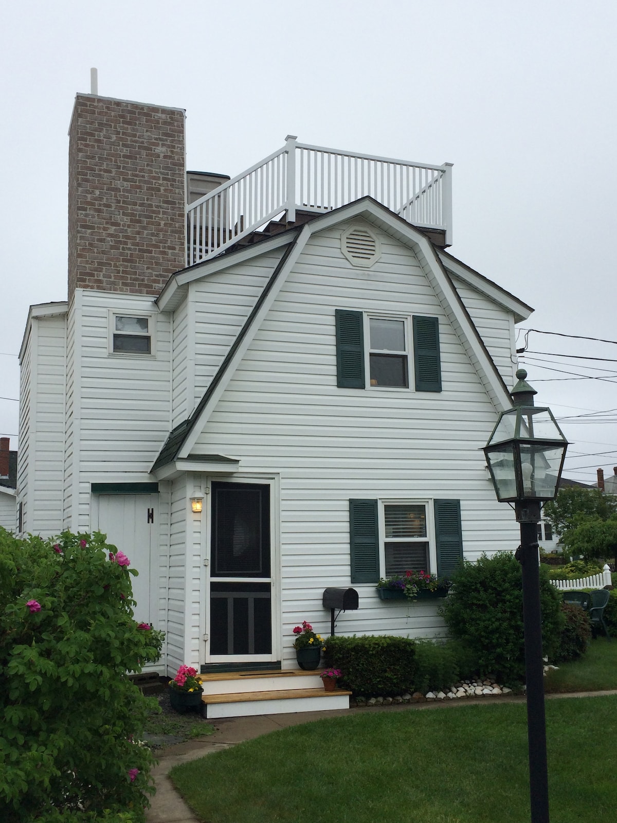 A two-story beach house with a white exterior and green shutters is shown. A rooftop deck is visible above the sloped roof, and a brick chimney adds visual interest. Lush greenery and flowers line the pathway leading to the entrance, enhancing the welcoming atmosphere.