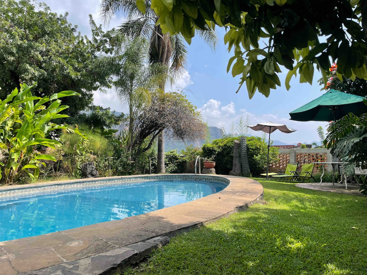 A heated swimming pool is surrounded by lush greenery, including palm trees and exotic plants. Sun loungers and an umbrella are arranged on the lawn beside the pool, with views of the mountains visible in the background under a clear blue sky.