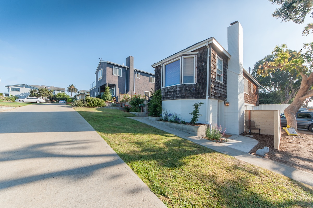A charming, two-story house is visible on the left, featuring a mix of dark wood siding and white trim. Adjacent to it, a single-story home is positioned, accompanied by a driveway that leads to a small garden area with greenery. The scene is bathed in soft morning light.