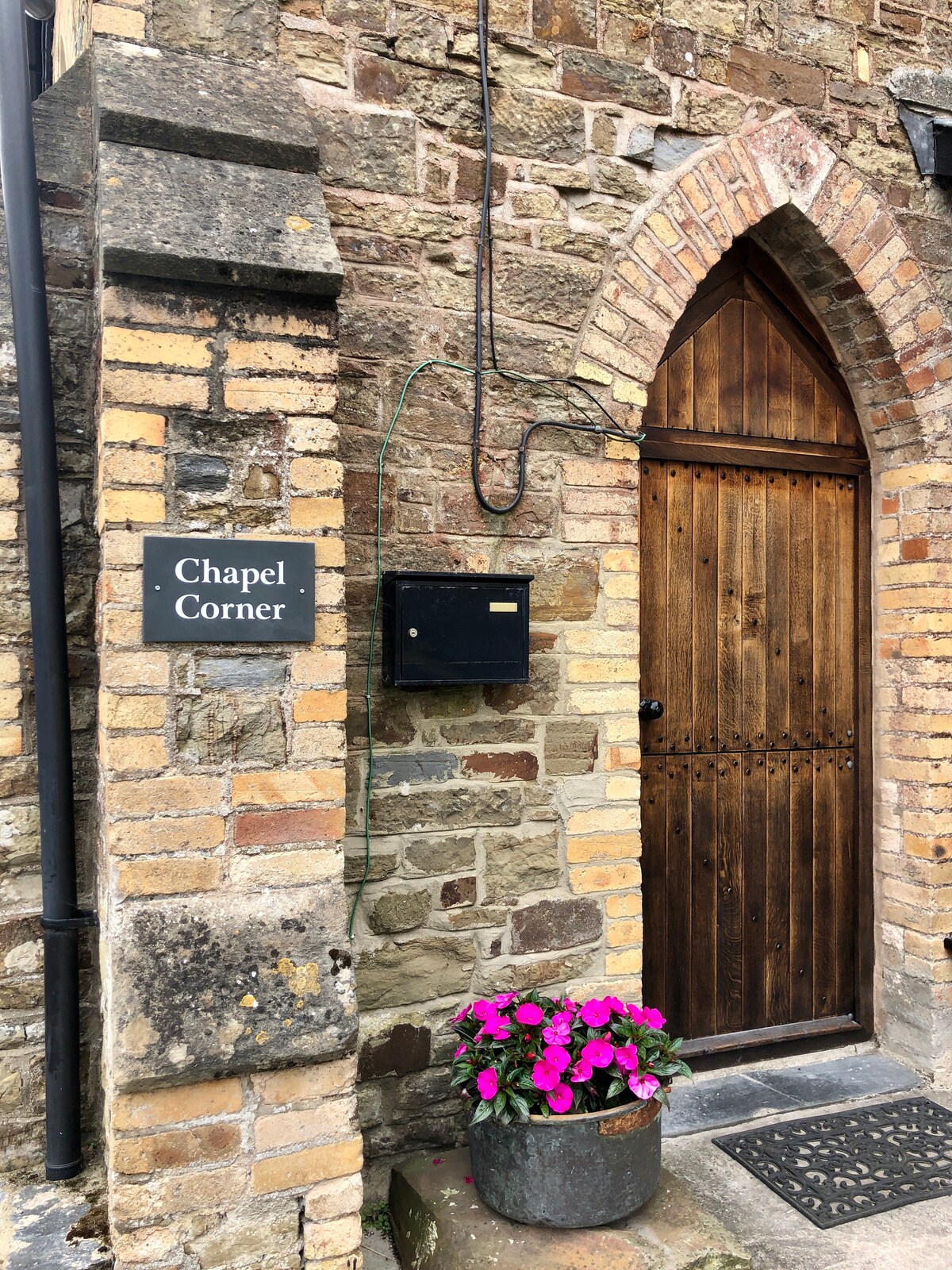 The entrance to Chapel Corner is framed by rustic stone walls, showcasing a wooden door with an arched top. A metal mailbox is mounted beside the door, and a planter with vibrant pink flowers adds a welcoming touch to the entryway.