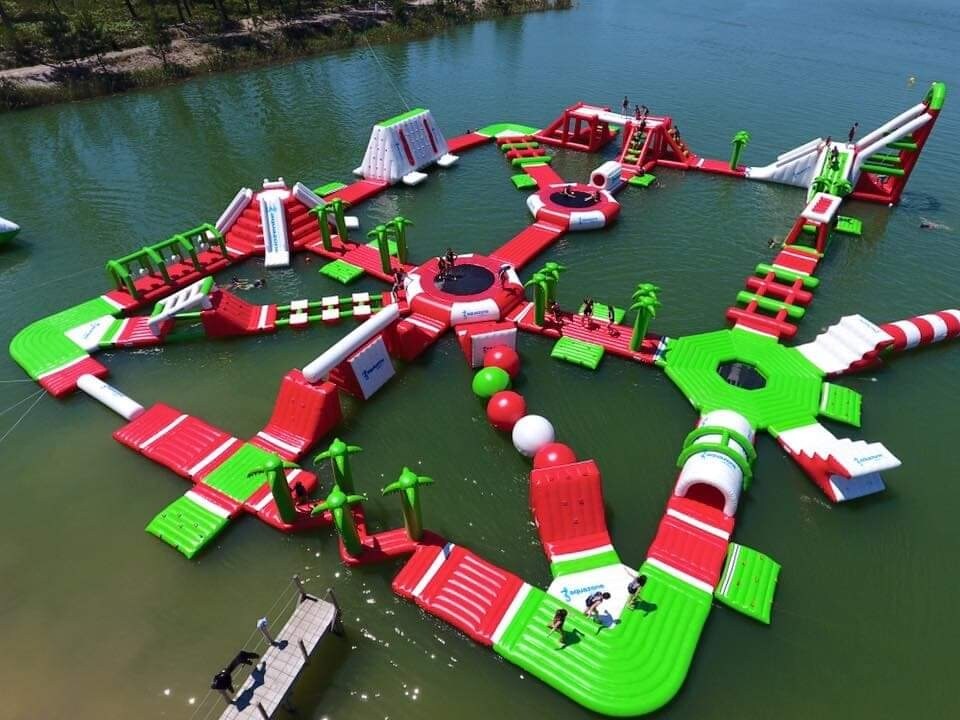 An expansive outdoor water park features bright red and green inflatable structures arranged in a star pattern. Various slides, trampolines, and climbing areas are visible, surrounded by calm water. Palm trees are scattered throughout, adding a playful touch to the vibrant environment.