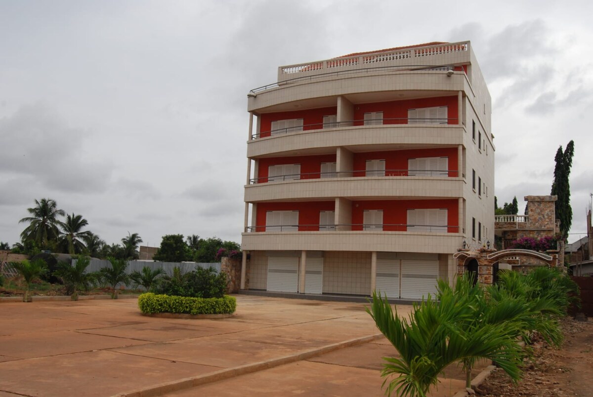 A modern three-story building is shown, featuring balconies on each level and large windows. The surrounding area is landscaped with green plants and palms, providing a calm environment. A spacious paved area in front allows for parking or outdoor use.