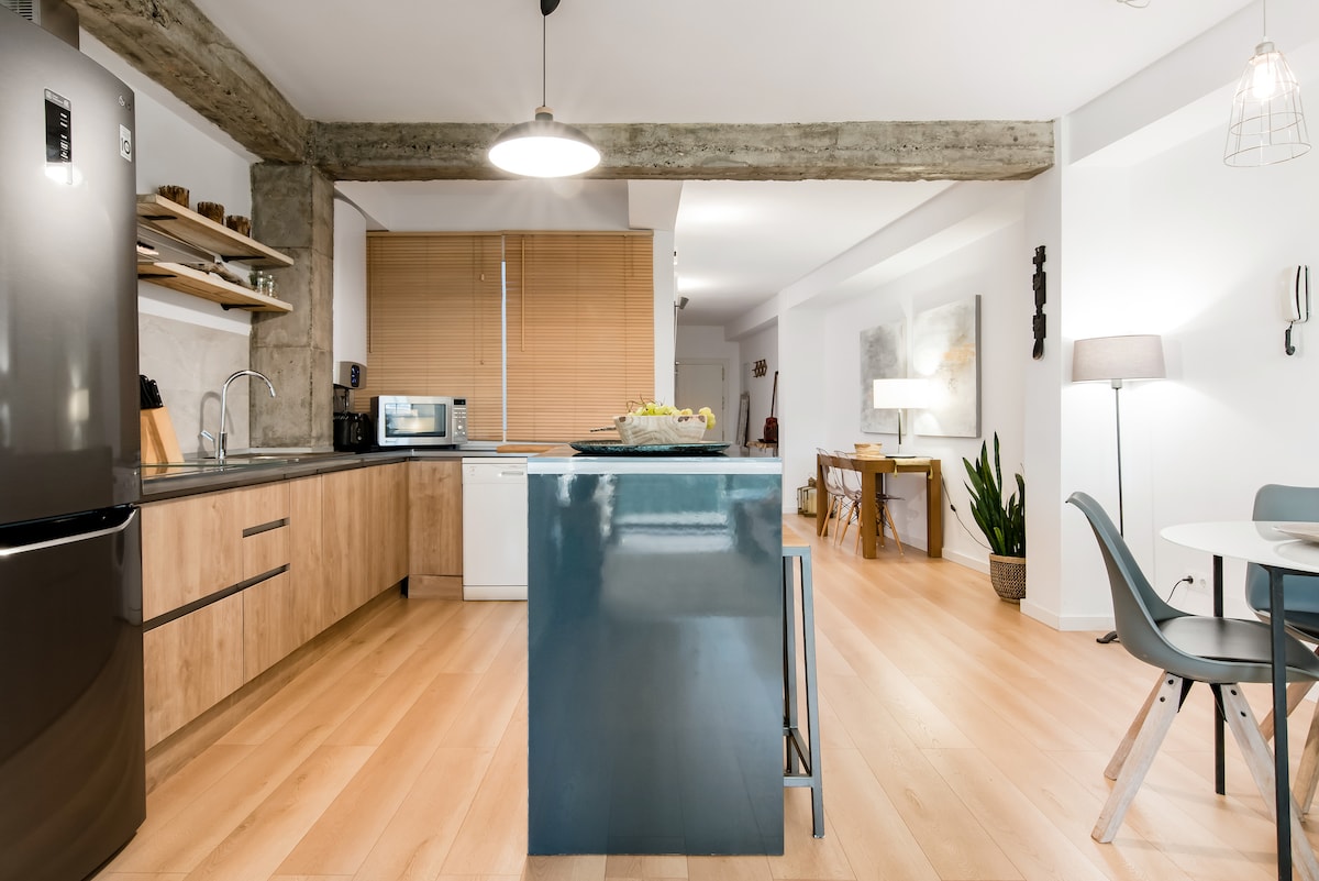 A modern kitchen features natural wood cabinetry and a sleek, dark island. The open layout is illuminated by overhead lighting, with a dining area visible in the background. Shelves display minimalistic decor, and sunlight filters in through the large window dressed with wooden blinds.