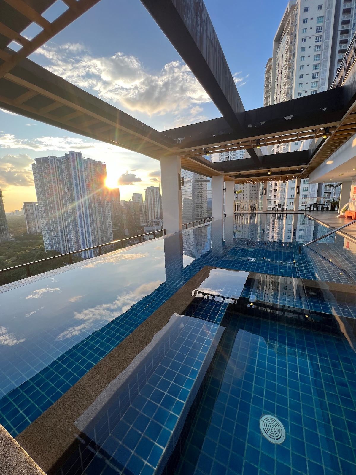 An infinity pool is framed by a spacious rooftop terrace, reflecting the colors of the sunset and surrounding skyline. The water surface showcases vibrant blue tiles, while modern architectural lines create a stylish setting for relaxation and enjoyment.