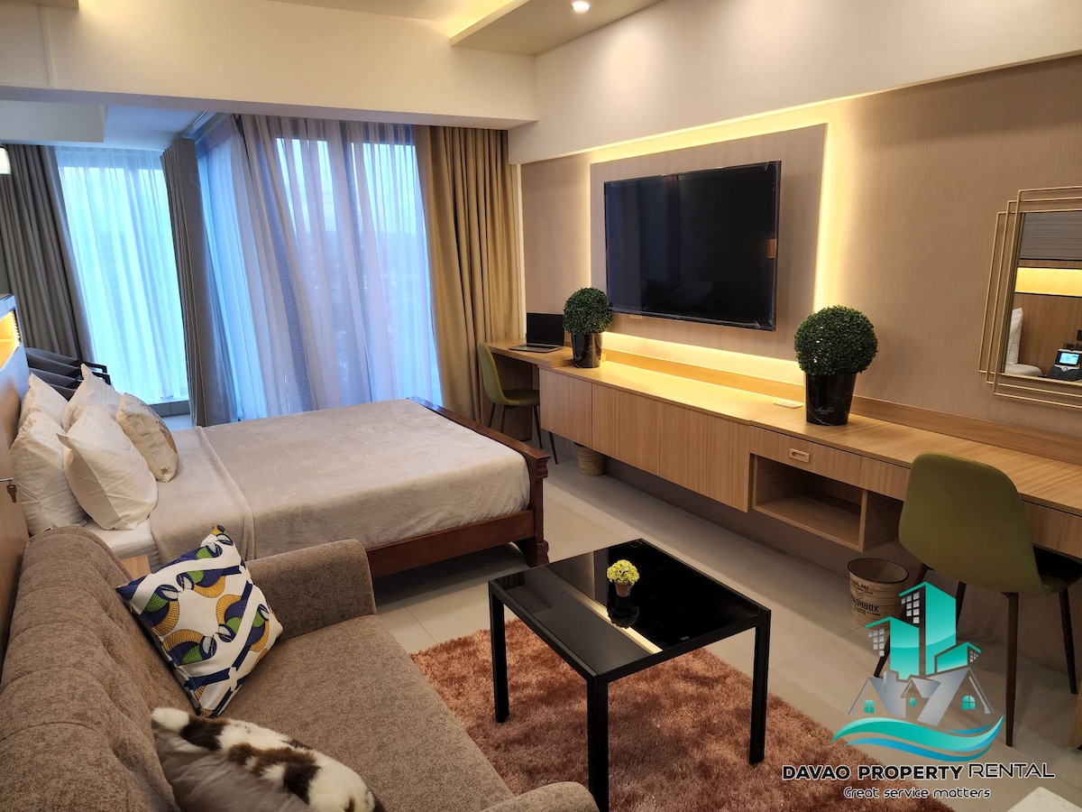 A modern studio unit is furnished with a spacious bed and a cozy seating area. The room features large windows allowing natural light to fill the space. A sleek television is mounted on the wall above a stylish console, complemented by decorative plants.