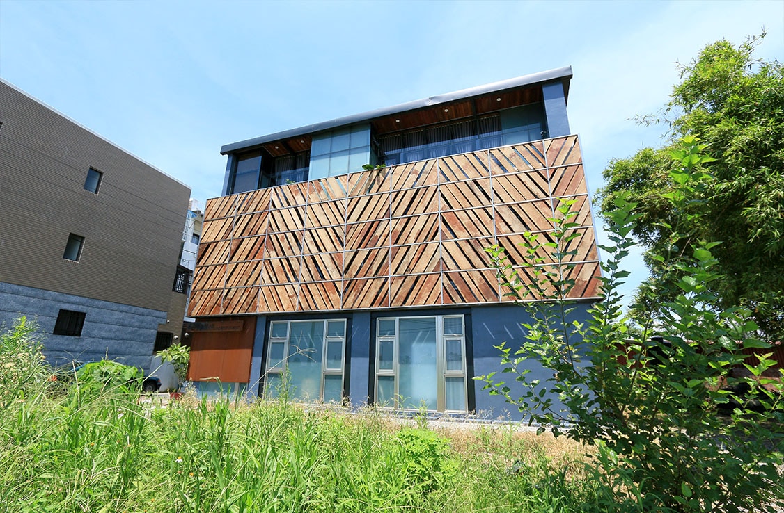 The exterior of a multi-story building displays a striking wooden facade with a geometric pattern. Large windows are visible on the ground and upper levels, allowing for natural light. Surrounding greenery enhances the building's presence against a clear blue sky.