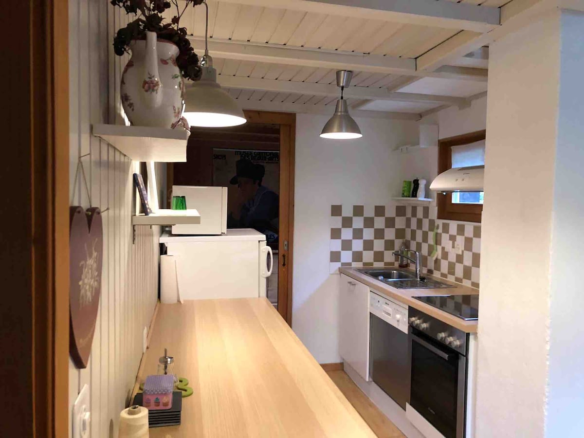 A well-equipped kitchen features modern appliances, including a refrigerator, dishwasher, and oven. Light wood countertops and tiled backsplash complement the clean, functional design. Pendant lights hang above, creating a bright workspace with ample natural light from a nearby window.