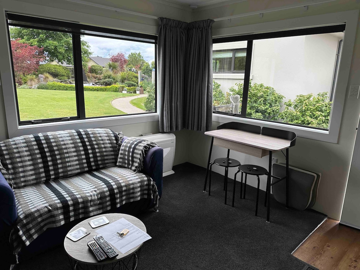 A cozy lounge area features a dark blue sofa draped with a patterned blanket. A small table is accompanied by two black chairs, positioned near large windows offering views of the surrounding greenery and pathway. Natural light brightens the space.