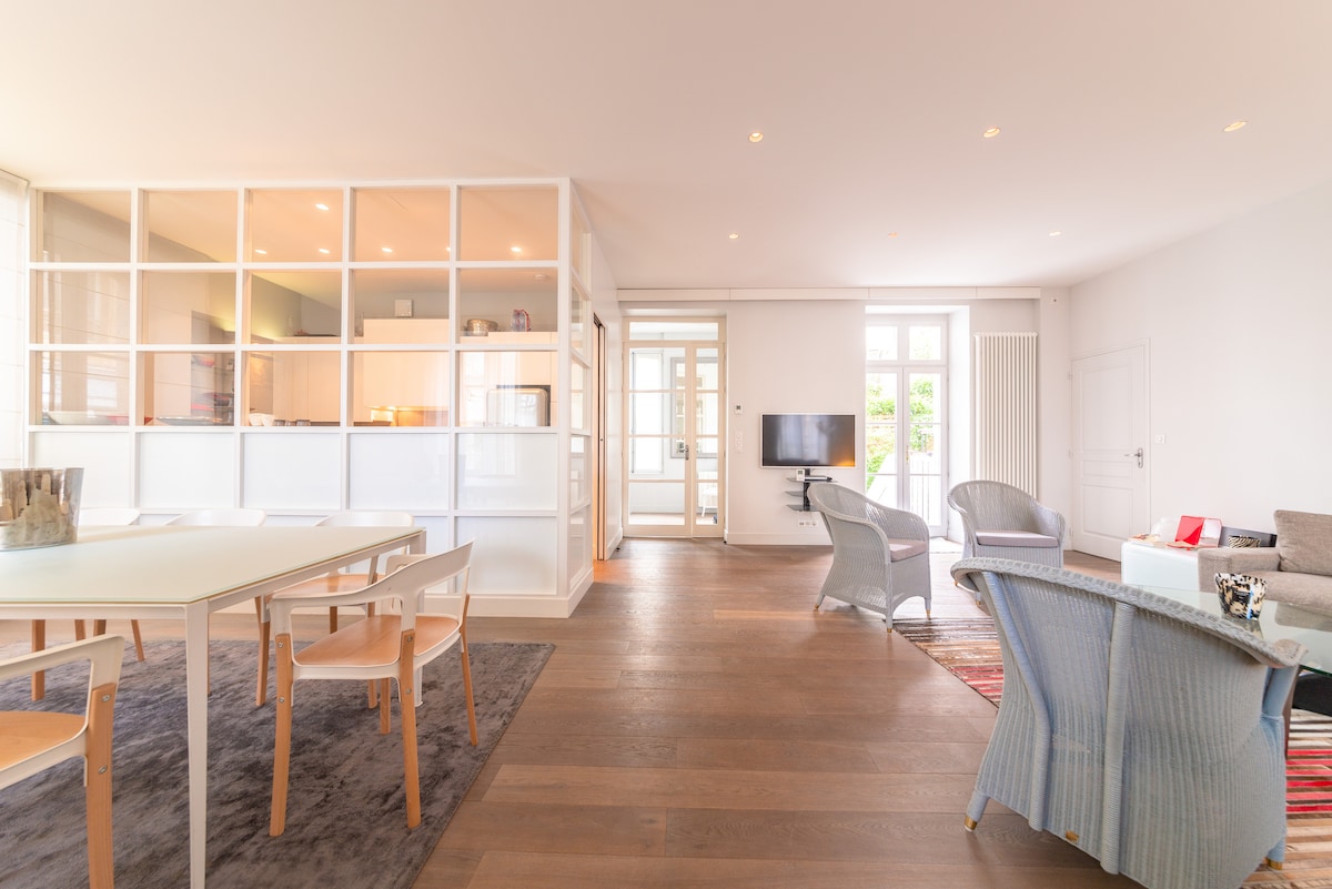 A bright and airy living space is illuminated by natural light, featuring a spacious dining table surrounded by light wooden chairs. A cozy seating area is visible with a soft gray couch and armchairs. The area connects seamlessly to a kitchen, enhancing the open layout.