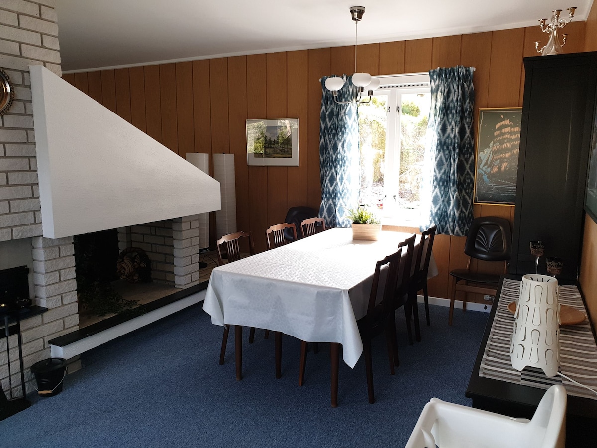 An inviting dining area features a large table with seating for eight, surrounded by wooden chairs. A stone fireplace adds to the warmth of the space, complemented by patterned curtains framing the window. Soft blue carpet covers the floor, enhancing the room's cozy atmosphere.