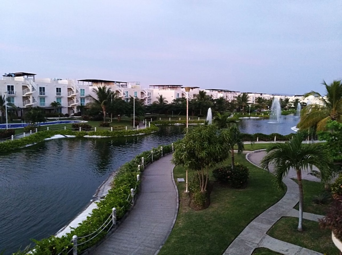 A serene view showcases well-maintained pathways bordered by lush greenery, winding along a tranquil artificial lake. Fountain jets create gentle splashes in the water. Modern buildings with balconies are visible in the background, framed by palm trees under a soft evening sky.