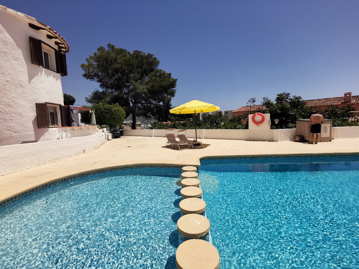 A communal pool area is depicted, featuring a clear blue pool surrounded by tiled decking. Two sun loungers with an umbrella provide shaded seating. Lush greenery can be seen in the background, along with a nearby building that enhances the tranquil setting.
