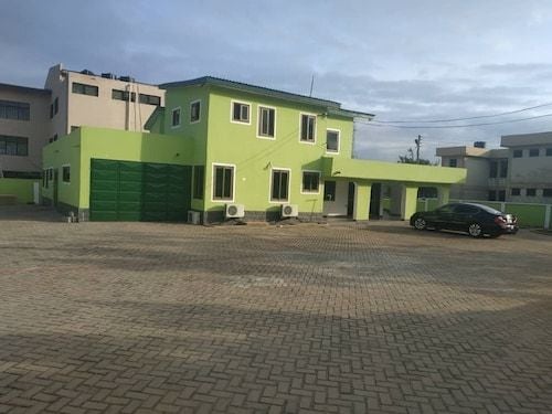 A two-story building features a vibrant green exterior with multiple windows and air conditioning units. A spacious driveway accommodates several vehicles, and an expansive parking area is visible in the foreground. The surroundings include other structures and clear skies.