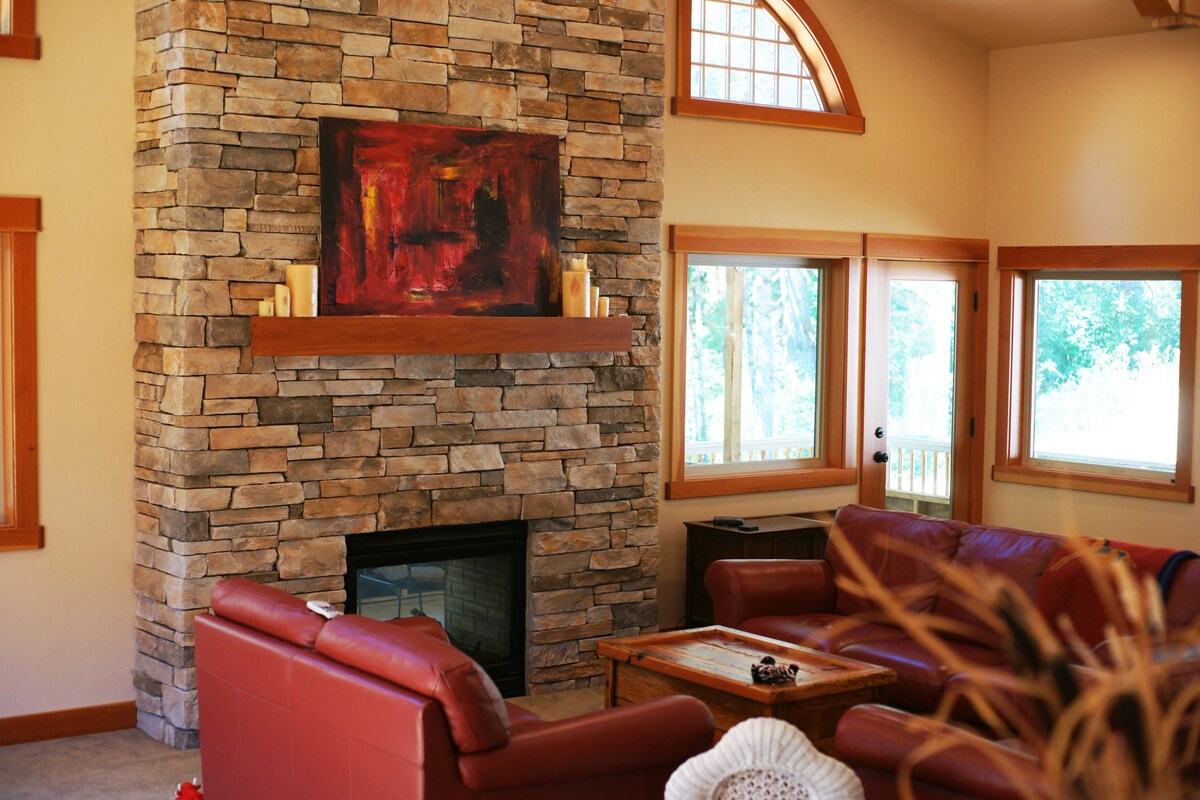 A warm living area features a stone fireplace as a focal point, complemented by a wooden mantel. Large windows allow natural light to illuminate the space, which includes a comfortable seating arrangement with deep red sofas and a wooden coffee table.