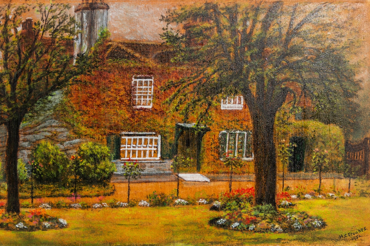 A traditional Georgian house is surrounded by trees and a well-maintained garden. The front entrance is framed by green shutters, and there are various shrubs and flowers lining the pathway, adding a touch of color to the scene.