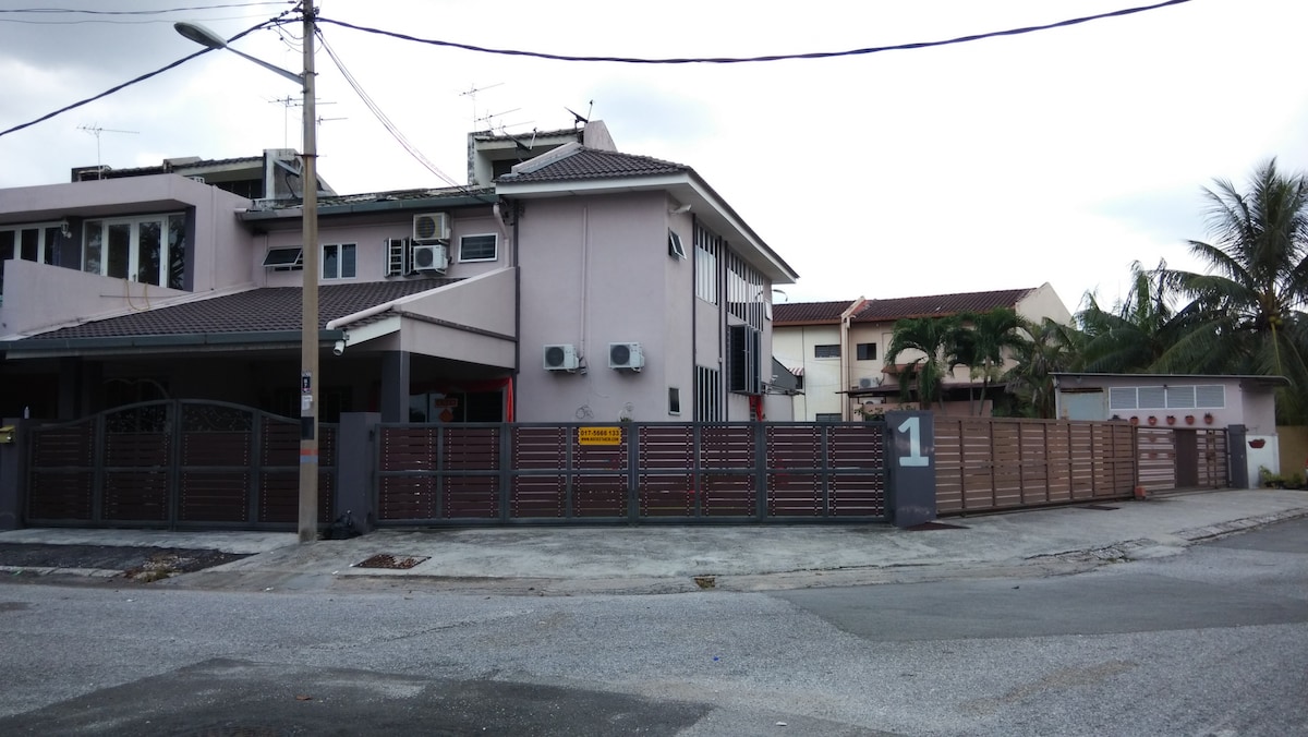 The exterior view of the guest house is presented, featuring a gated entrance with a numbered marker. The structure includes multiple air conditioning units, and greenery is visible in the surrounding area. Paved pathways and a residential neighborhood atmosphere are evident.