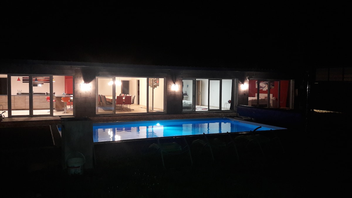 A contemporary villa is illuminated at night, showcasing a tranquil pool in vibrant blue tones. Expansive glass doors provide glimpses into the inviting indoor spaces, where soft lighting creates a welcoming ambiance. Outdoor seating is visible beside the pool, enhancing the relaxing atmosphere.