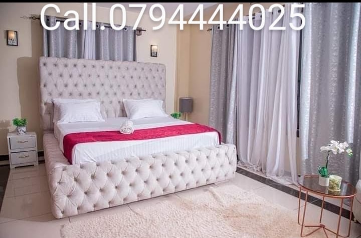 A spacious sleeping area is furnished with a large tufted bed featuring a soft cream upholstery and red accents. Towels are artfully arranged on the bed. Natural light filters through sheer curtains, illuminating the room, complemented by a small round table and decorative plants.