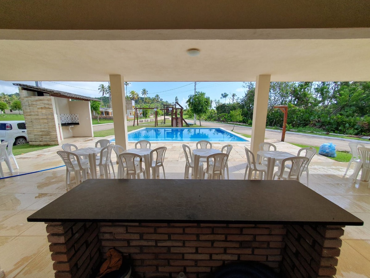 A spacious patio is visible, with a stone bar counter and several white plastic chairs arranged around a table. In the background, a swimming pool glistens, surrounded by lush greenery and a well-maintained lawn. Natural light fills the area, creating an open, inviting space.