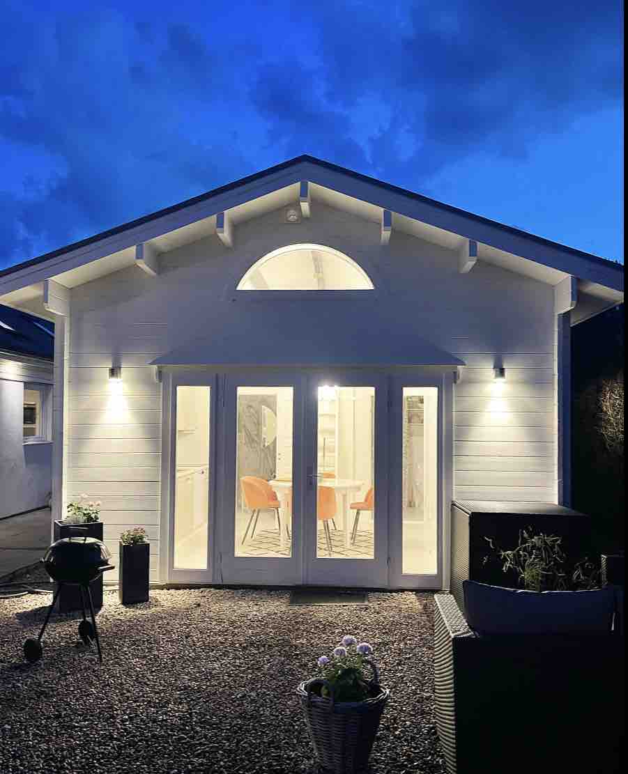 The exterior of the guesthouse is illuminated at dusk, featuring large glass doors that provide a view into the cozy interior. A grill is positioned nearby, surrounded by decorative planters. The roof displays an arched window, adding a charming architectural detail.