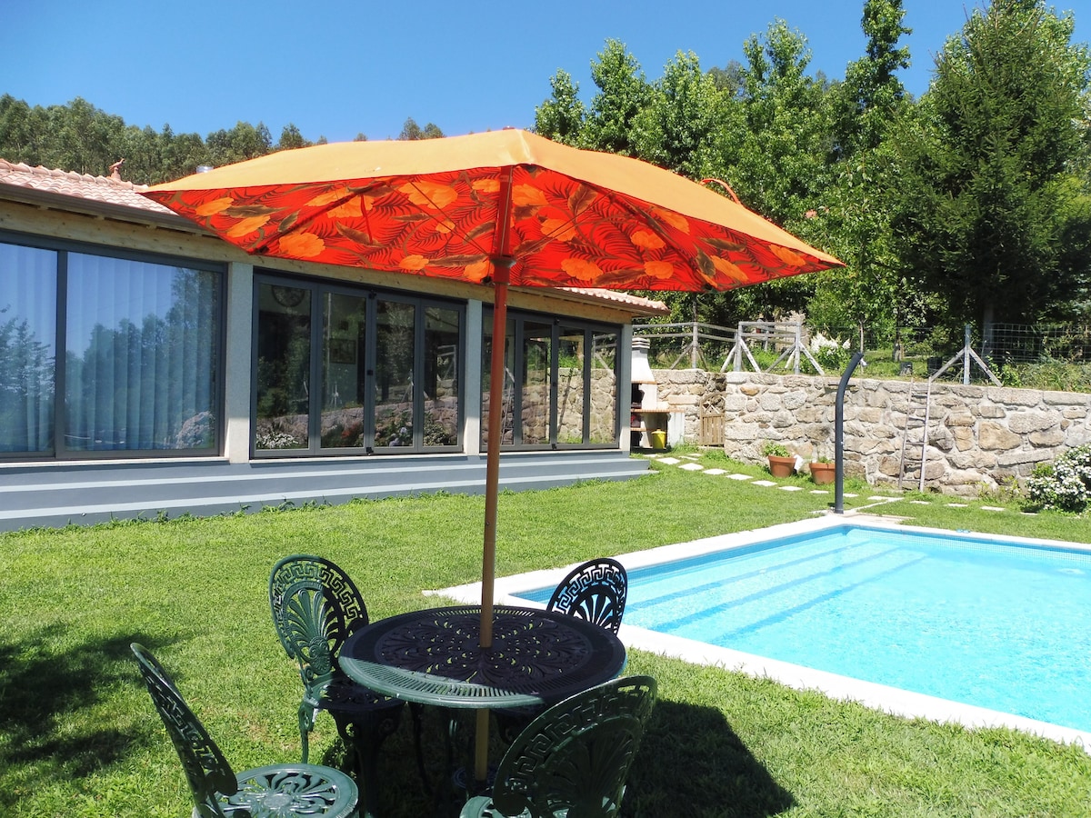 An orange umbrella provides shade over a round table surrounded by four green chairs, set on lush grass. A rectangular pool is situated nearby, framed by manicured greenery and a stone wall, while large glass windows of the house reflect the outdoor scenery.