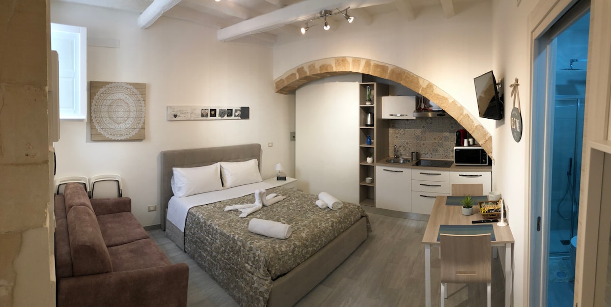 The image shows a cozy monolocale with a spacious bed, a comfortable sofa, and a dining table set for two. Modern kitchen amenities are visible, and a soft color palette creates a serene environment. Arched stone details add a touch of character to the space.