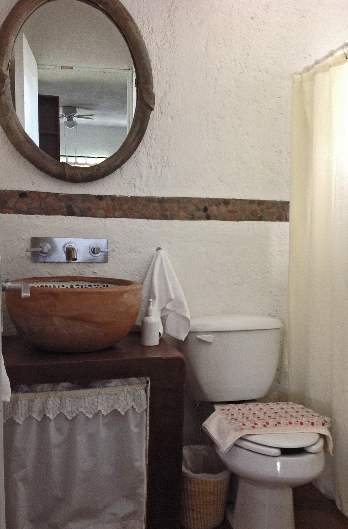 The bathroom features a rustic design with a large, round, stone sink mounted on a wooden base. A mirror with a wooden frame hangs above the sink. A shower with a curtain is visible, along with a toilet and a small wastebasket.