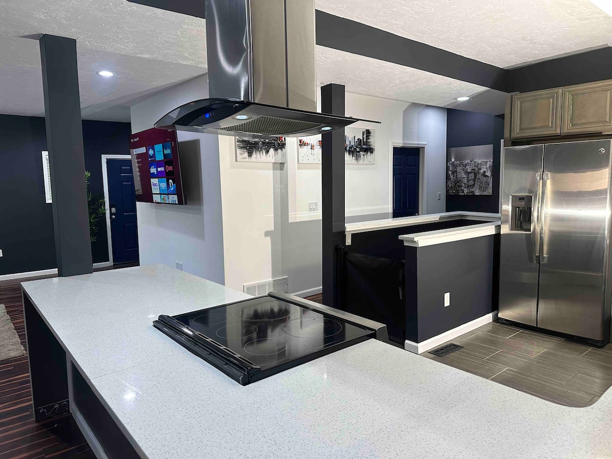 The kitchen showcases a modern design with a central island featuring an induction cooktop. A stainless steel refrigerator stands beside warm wooden cabinets. A flat-screen television is mounted on the wall, while sleek countertops and a dark accent wall enhance the contemporary aesthetic.
