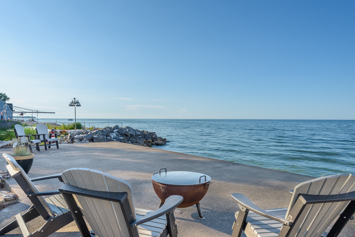 Adirondack chairs are arranged around a fire pit on a smooth stone patio, facing the calm waters of Lake Ontario. The shoreline is lined with rocks, and nearby greenery adds natural beauty to the serene waterfront setting under a clear blue sky.