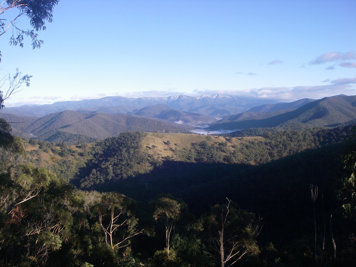 A wide view of rolling mountains stretches into the distance, with varying shades of green and blue creating a serene landscape. Snow-capped peaks are visible in the background, while a valley below reveals a hint of mist, enhancing the natural beauty of the scene.