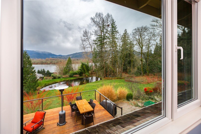 A scenic view from the window captures a spacious deck with outdoor seating and a dining table, surrounded by lush greenery. A small pond is visible in the background, along with trees and distant hills, all under a cloudy sky.