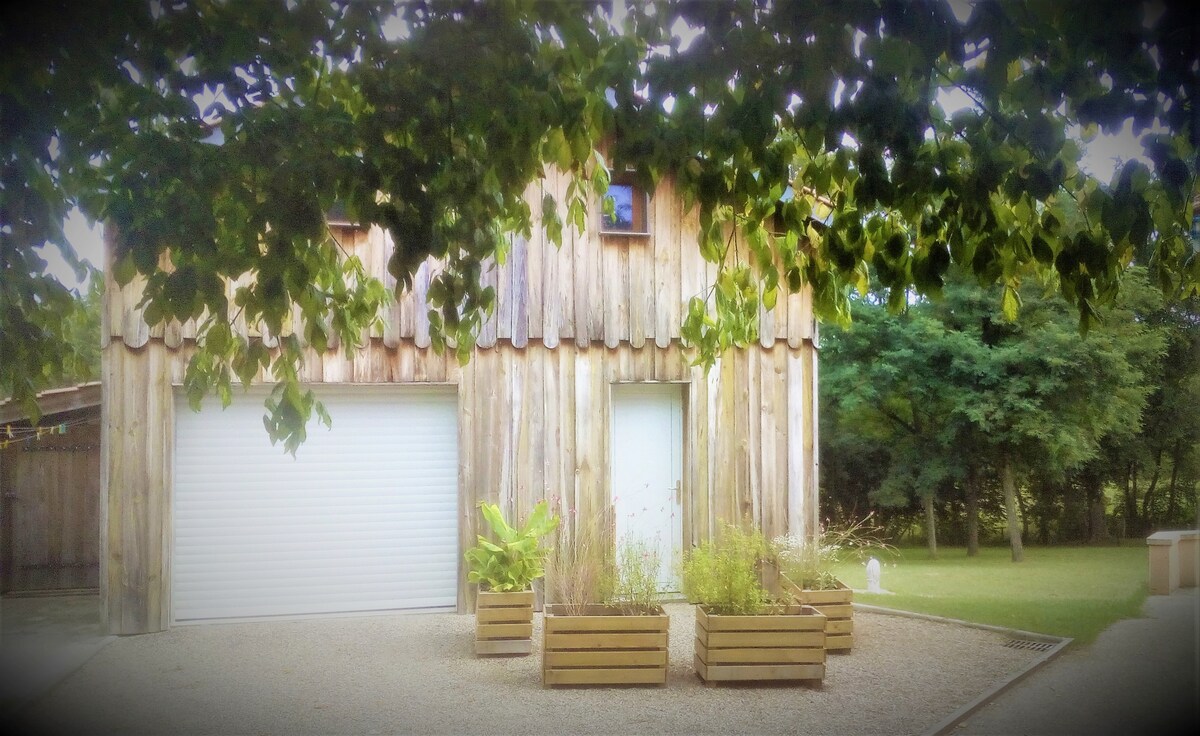 The exterior of the chalet showcases a wooden facade with a sloped roof. Two garage doors are visible beneath a canopy of leaves. Planters with greenery line the walkway, while a spacious lawn is seen in the background, framed by trees.