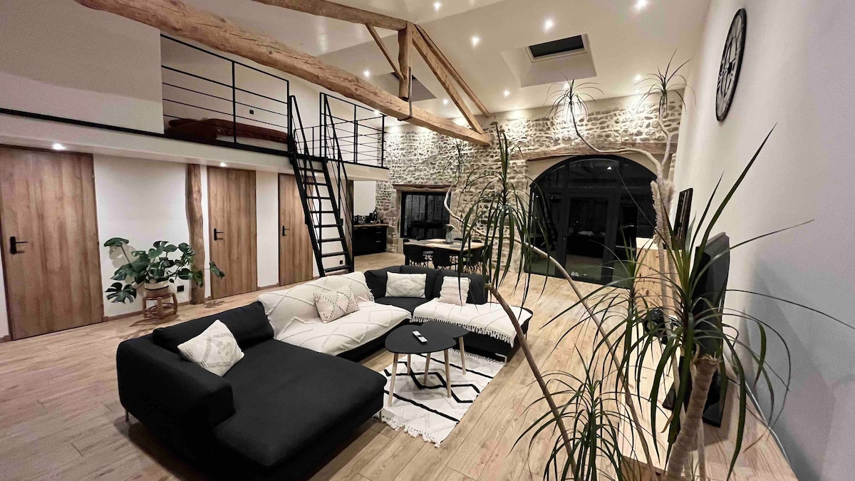 A spacious living area features a large sectional sofa with contrasting cushions, a low coffee table centered on a patterned rug, and floor-to-ceiling windows allowing natural light to fill the space. Exposed wooden beams and a ladder to a loft area add character to the room.