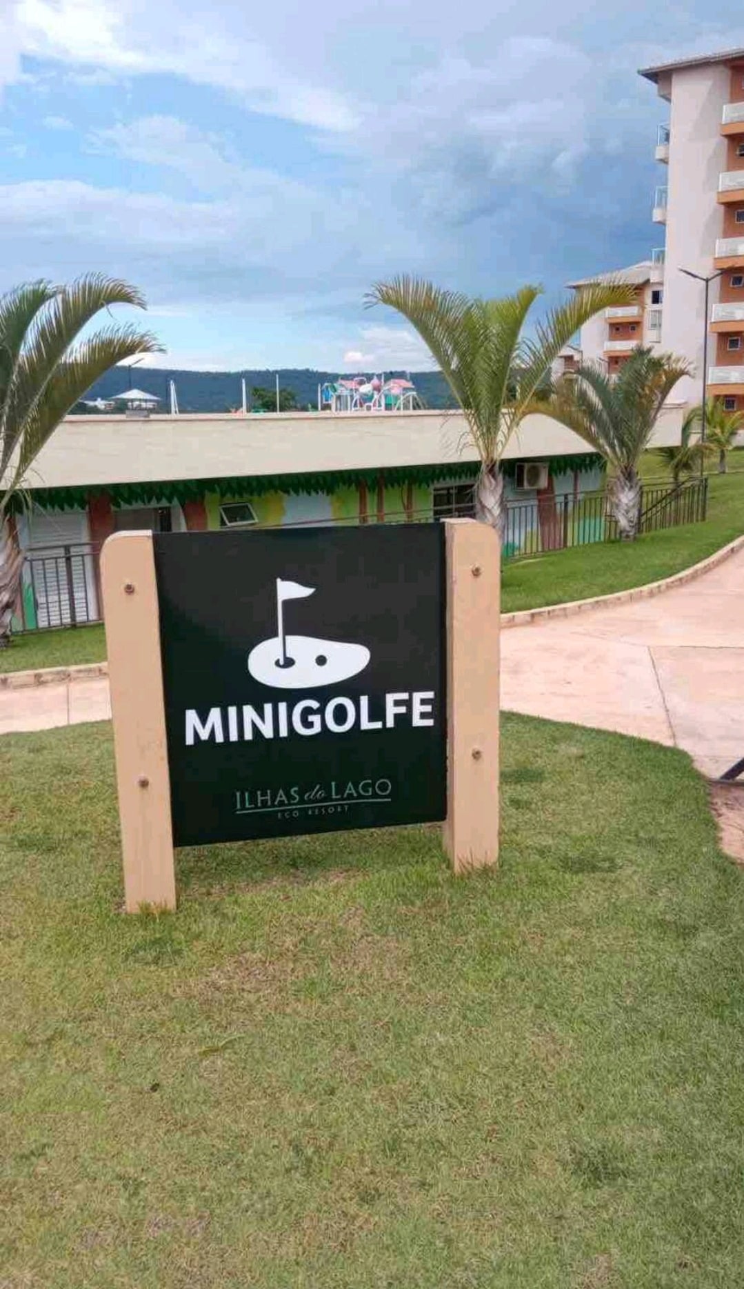 A sign designates the entrance to a mini-golf area, featuring a simple icon of a golf flag. Lush green grass surrounds the sign, and palm trees are visible in the background, suggesting a tropical atmosphere.