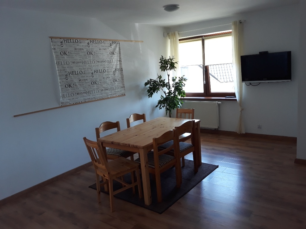 A dining area features a wooden table surrounded by four chairs, with a small plant positioned nearby. A wall hanging with text decorates the space, and a television is mounted on the wall opposite a large window, allowing natural light to enter.