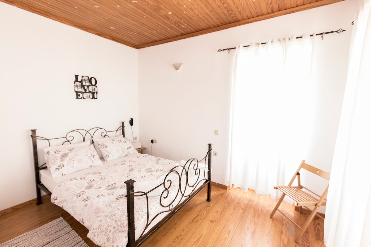 A cozy bedroom features a double bed with light-colored bedding and decorative pillows. Natural light streams through a large window, complemented by sheer white curtains. A wooden chair is positioned in the corner, and the ceiling showcases wooden planks, creating a warm atmosphere.