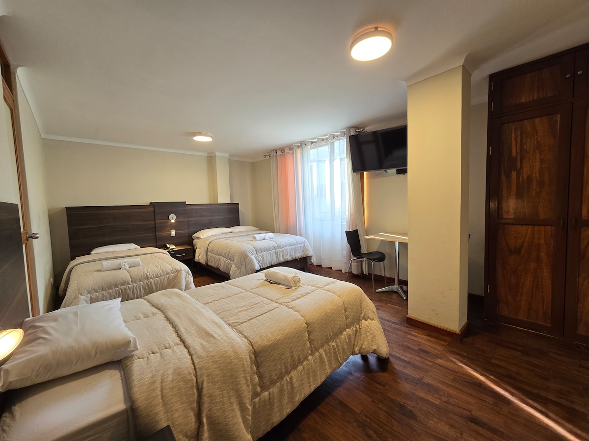 A spacious guest room with three beds arranged neatly, featuring light wood accents and soft bedding. Natural light enters through a corner window, complementing a flat-screen TV mounted on the wall. A desk and chair are positioned near a wardrobe, providing additional convenience.