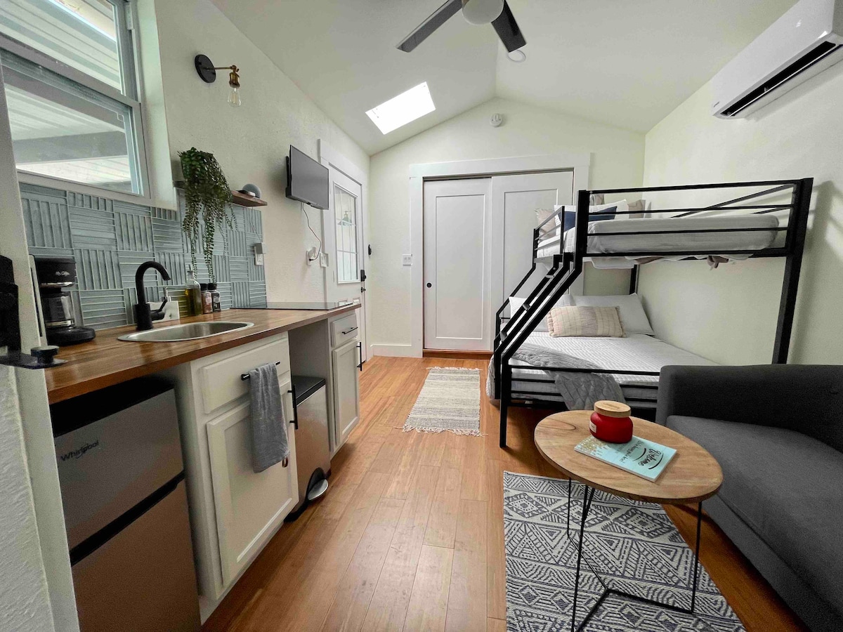 The compact living space includes a modern kitchenette with a small refrigerator and an electric stovetop. A cozy seating area features a dark sofa, while a bunk bed provides additional sleeping options in the background. Natural light streams in through a skylight.