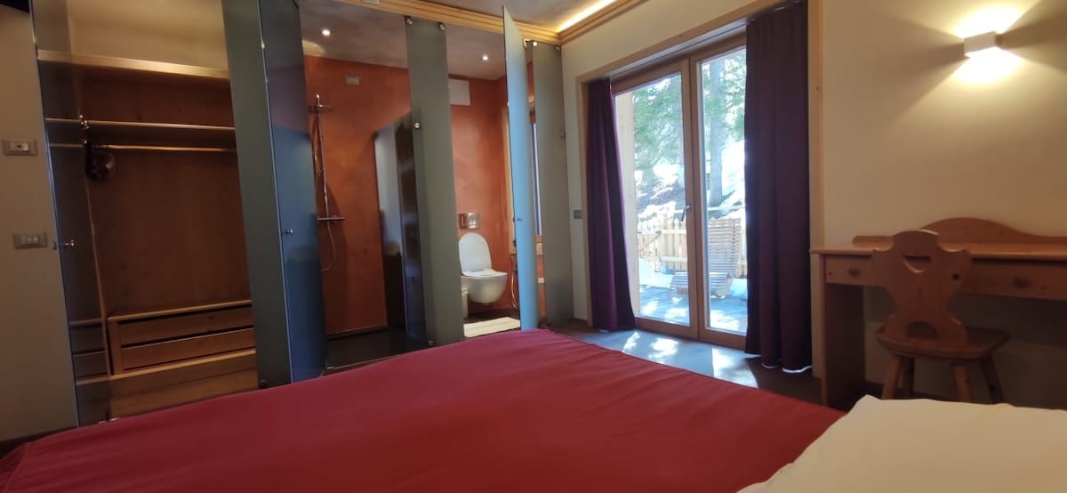 A spacious bedroom features a wooden headboard and warm-toned walls. A large window offers views of the outdoors, while a closet and a wooden chair are positioned nearby. Natural light fills the room, enhancing the cozy ambiance.