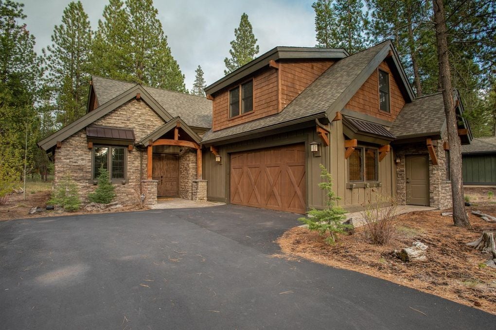 The exterior of a mountain modern home is showcased, featuring a blend of stone and wood materials. A paved driveway leads to a spacious garage with a large door. Tall pine trees and natural landscaping surround the home, contributing to a serene outdoor setting.