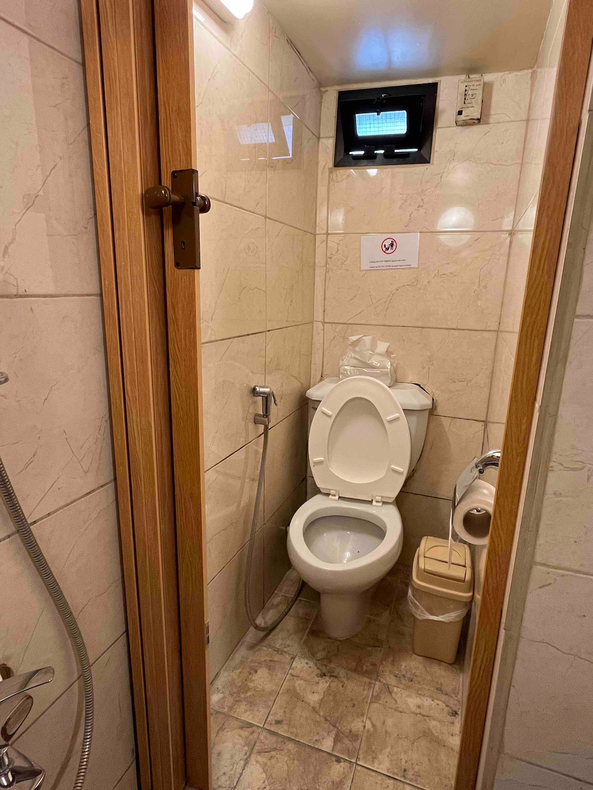 A compact bathroom features light-colored tiles and a modern toilet. The space includes a handheld showerhead and a small waste bin. A window near the ceiling provides ventilation, while a sign on the wall offers additional information.