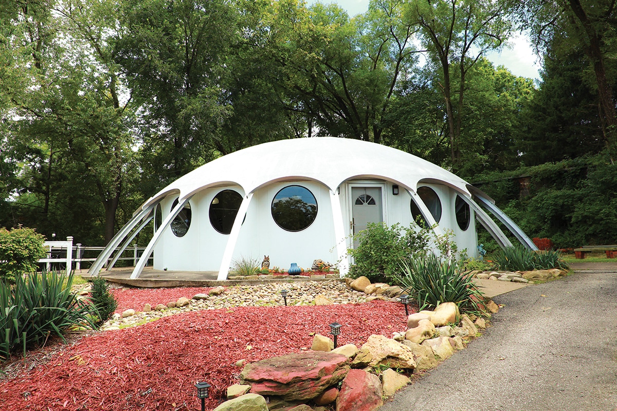 The exterior of the Yaca-Dome is visible, featuring a distinctive curved white structure with large circular windows. Surrounding the dome, a landscaped area includes red mulch, stones, and green shrubs, creating a harmonious blend with the natural wooded surroundings.